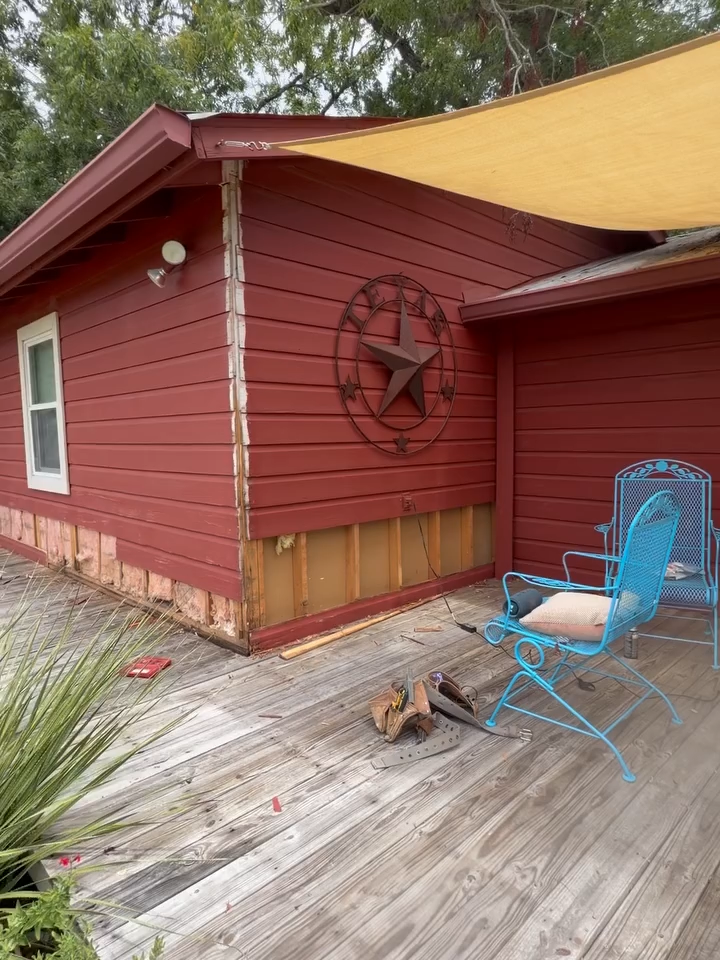 Red wooden cabin with Texas star decor, blue chair, and tan shade sail on a weathered deck.