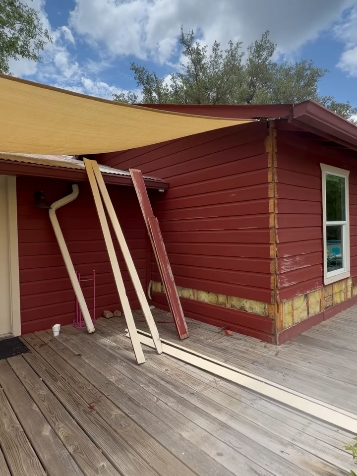Red house exterior with damaged awning, tilted support beams, and wooden deck under a cloudy sky