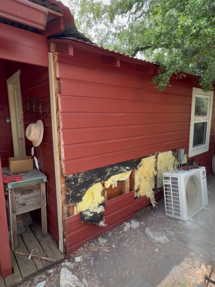Damaged red exterior wall with exposed insulation beside a porch and white plastic stools