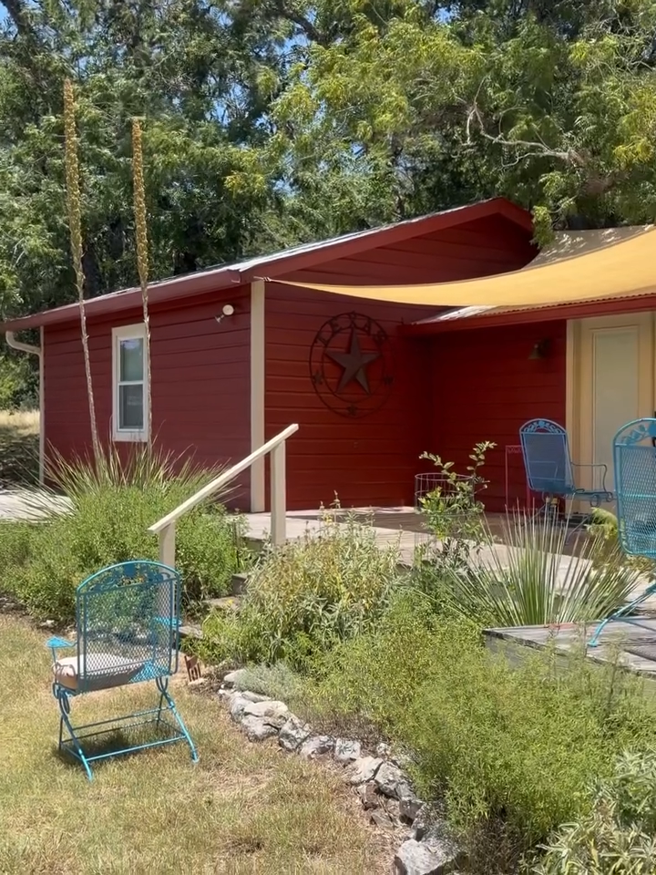 Red house with a small porch, blue chair, and overgrown yard with trees in the background.