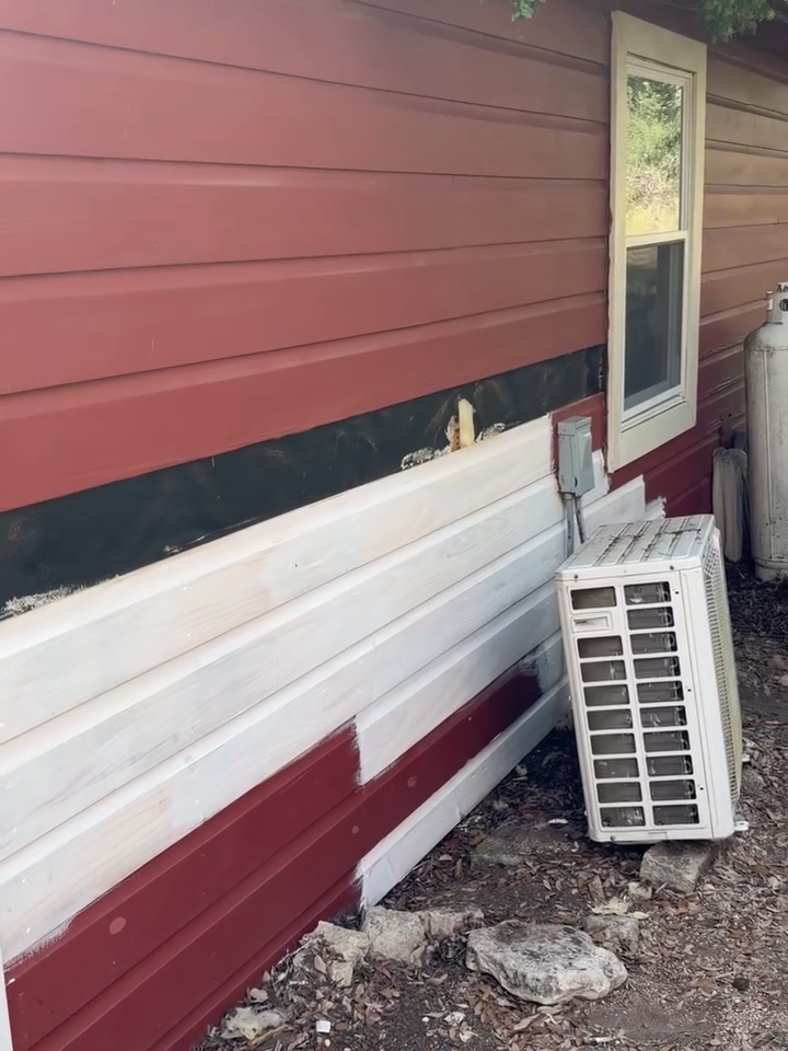 Damaged red and white house siding with a detached window AC unit leaning beside it.
