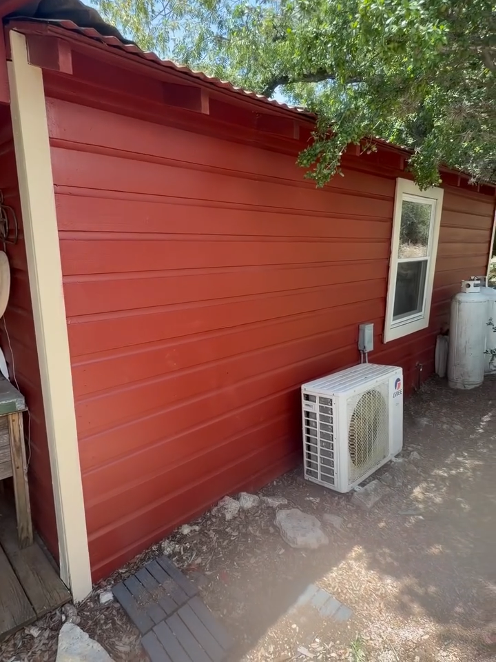 Red wooden exterior wall with a window and outdoor air conditioner unit beside a gravel path.