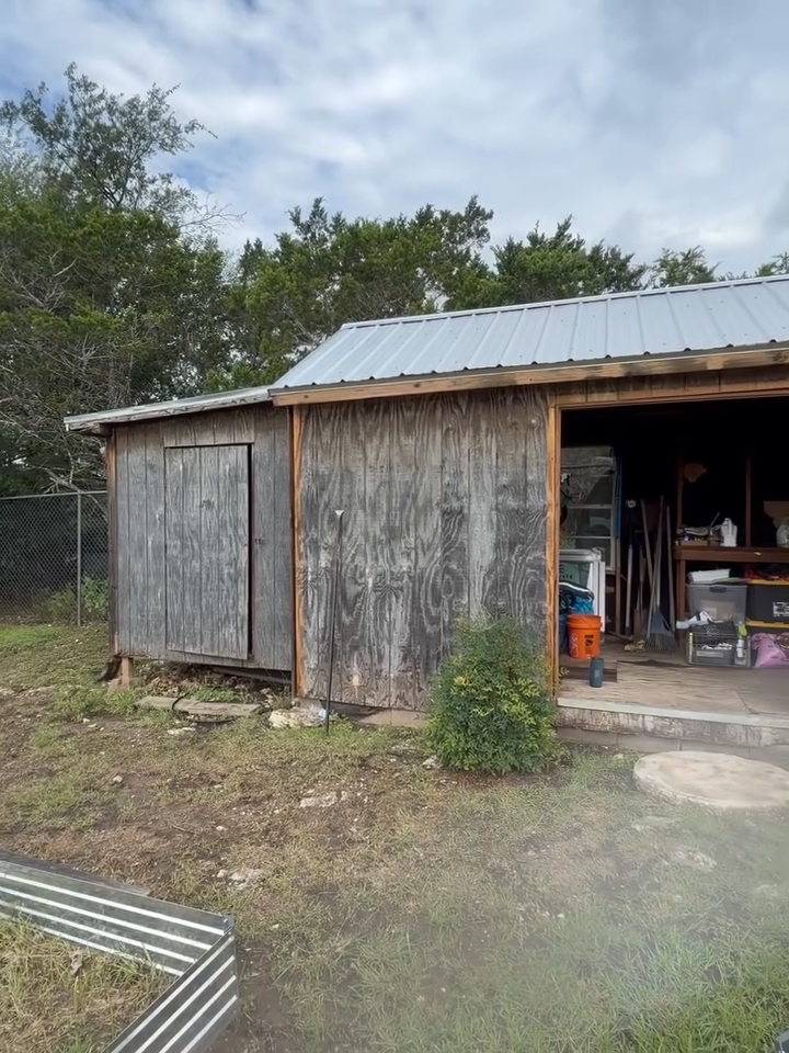 Old weathered shed with a metal roof, open doorway, and outdoor yard with tools and vegetation.
