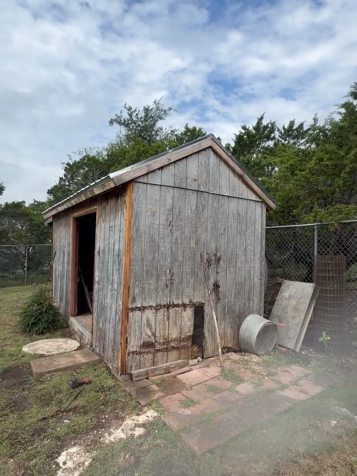 Weathered wooden shed with open doorway in a fenced yard, with a bucket and ramp beside it.