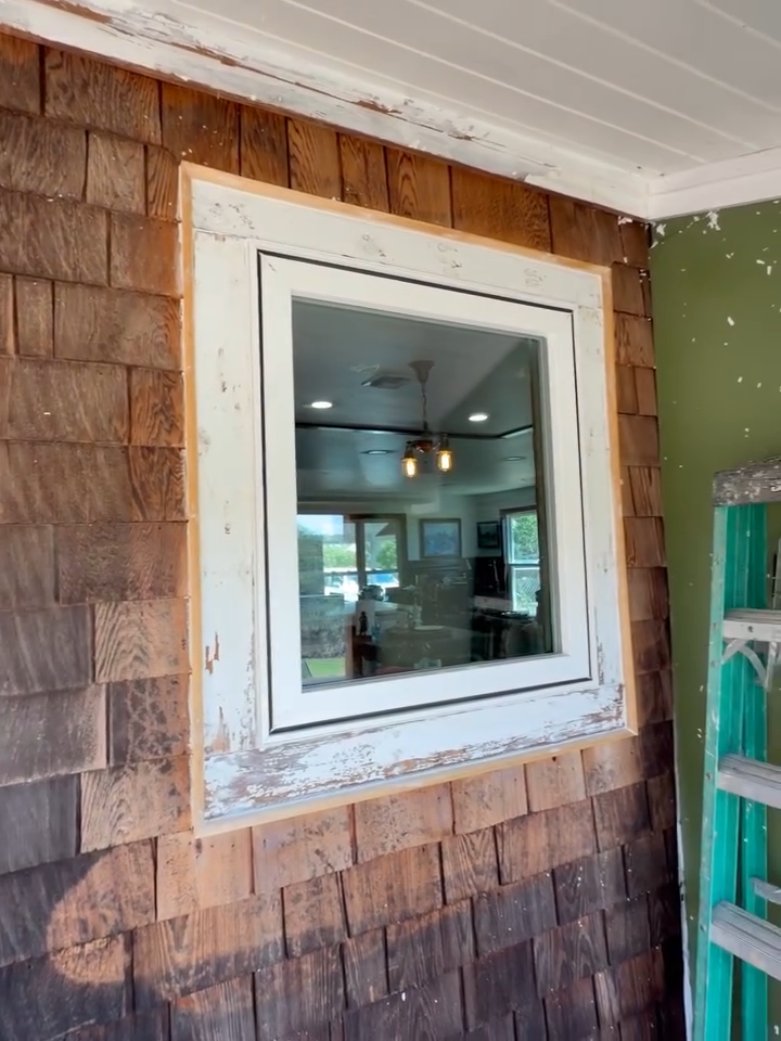 White-framed window in a weathered brown shingle exterior wall, with interior lights visible through the glass.
