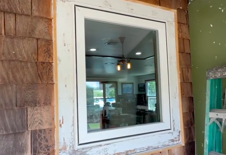White-framed interior window in a rustic wood exterior wall, showing a room with ceiling fan and windows inside
