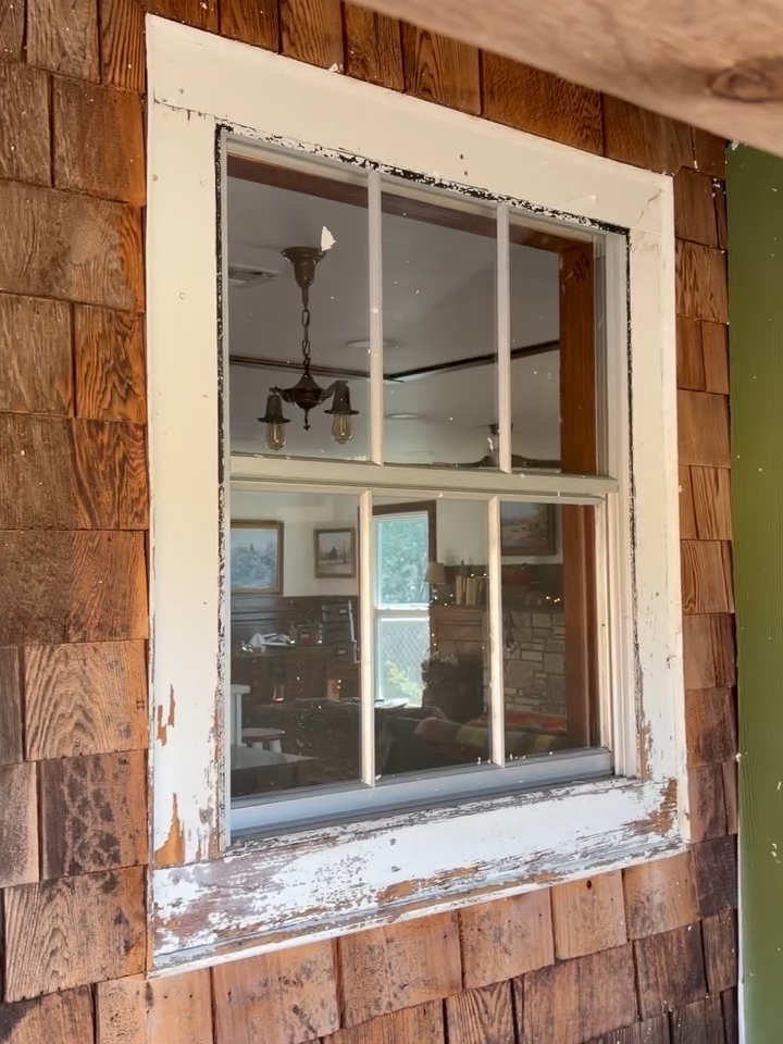 Weathered white-trimmed window set in brown shingle siding, showing a room interior with a ceiling fan.