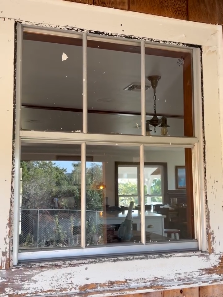 Old white-trim window with chipped paint reflecting an interior room and outdoor trees