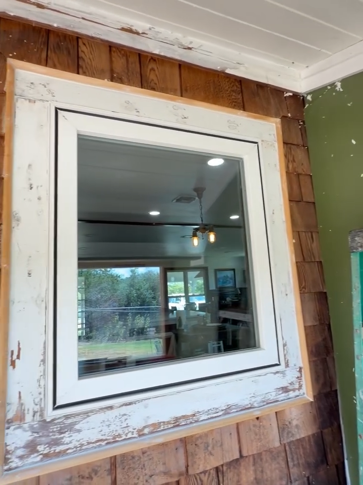 White-framed window in a wood-shingled wall with peeling paint, reflecting an indoor room and outdoor yard.