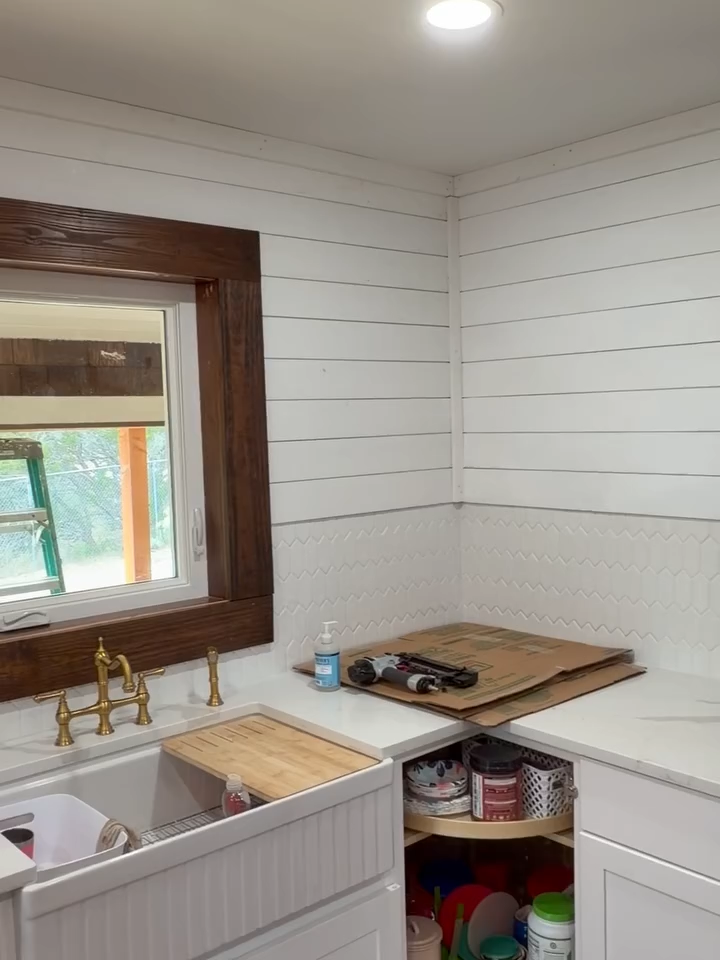Bright kitchen sink and counter with brass faucet, white shiplap walls, and a window over the sink