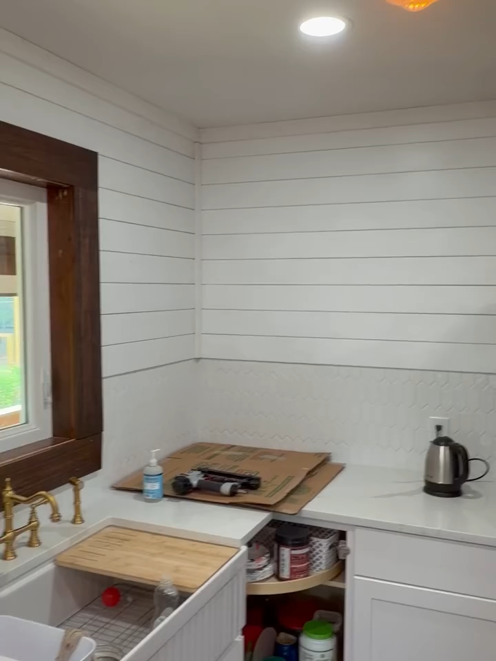 Small kitchen corner with white shiplap walls, countertop, sink, window, kettle, and a wooden cart with items.