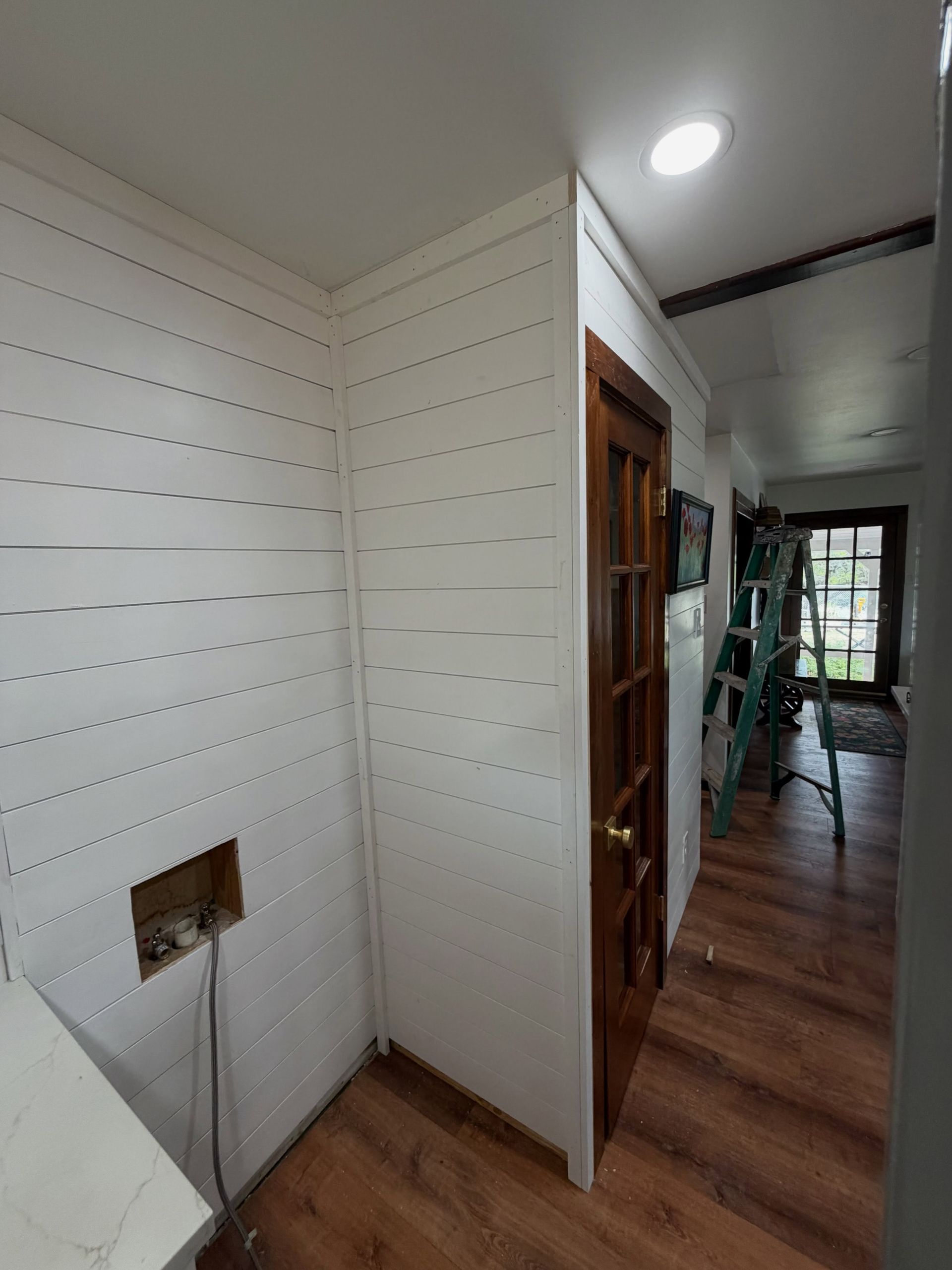 Narrow hallway with white panel walls, wood floor, and a wooden door leading to a brighter room.