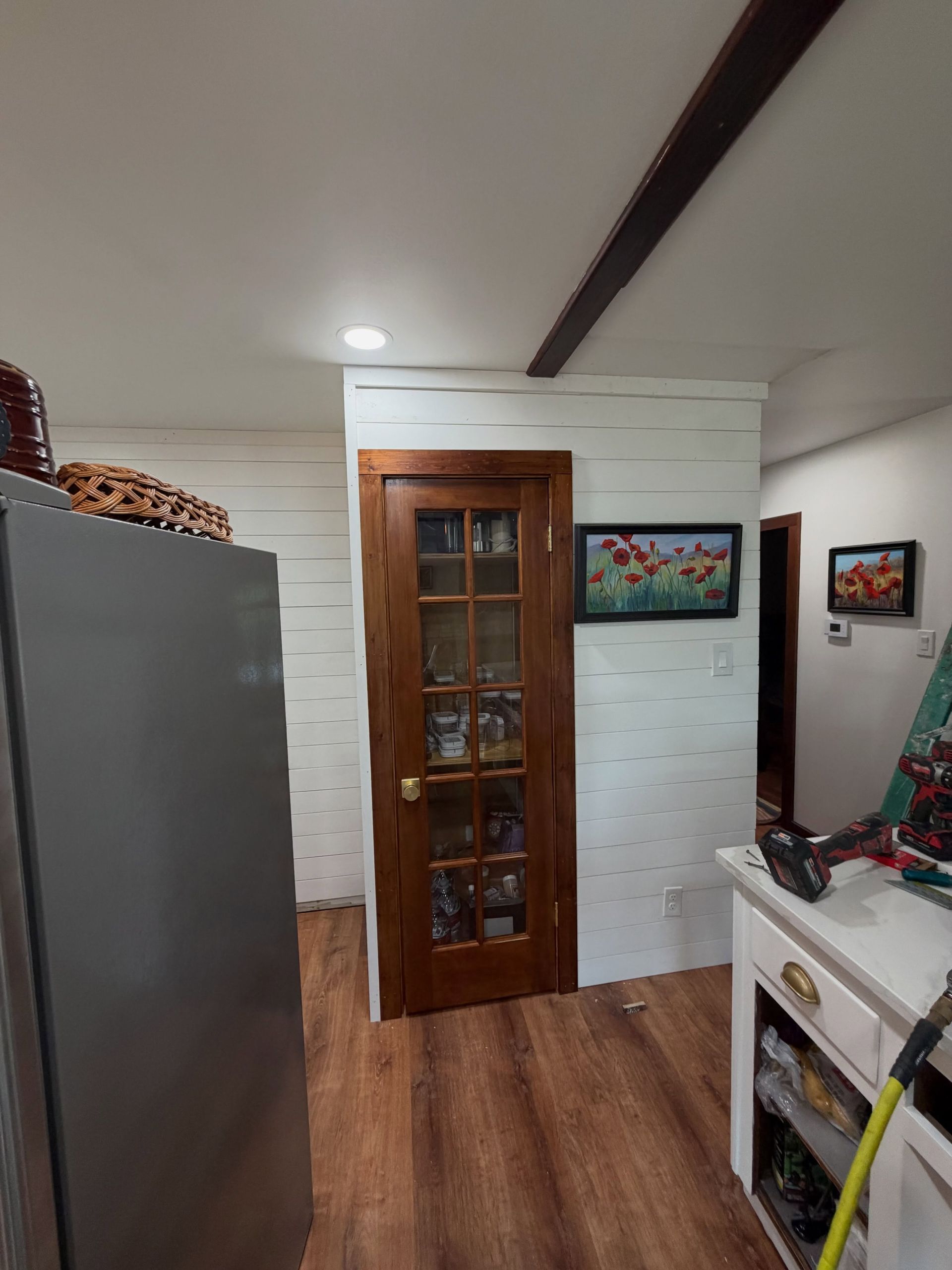 Narrow kitchen with white walls, wood floor, gray fridge, wooden cabinet door, and small countertop items