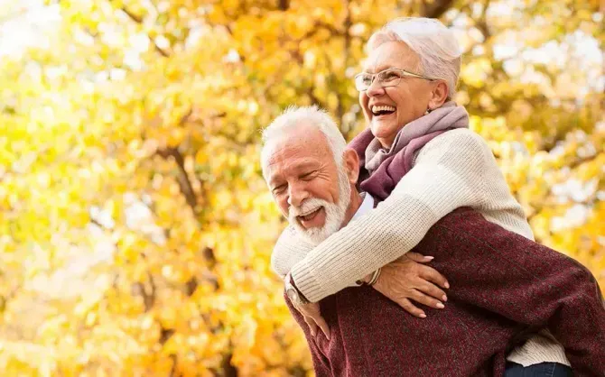 A man is giving an older woman a piggyback ride in a park.