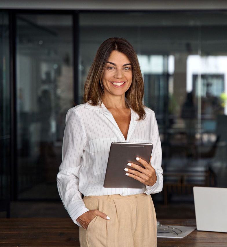 A woman is holding a tablet and smiling while standing in front of a laptop.