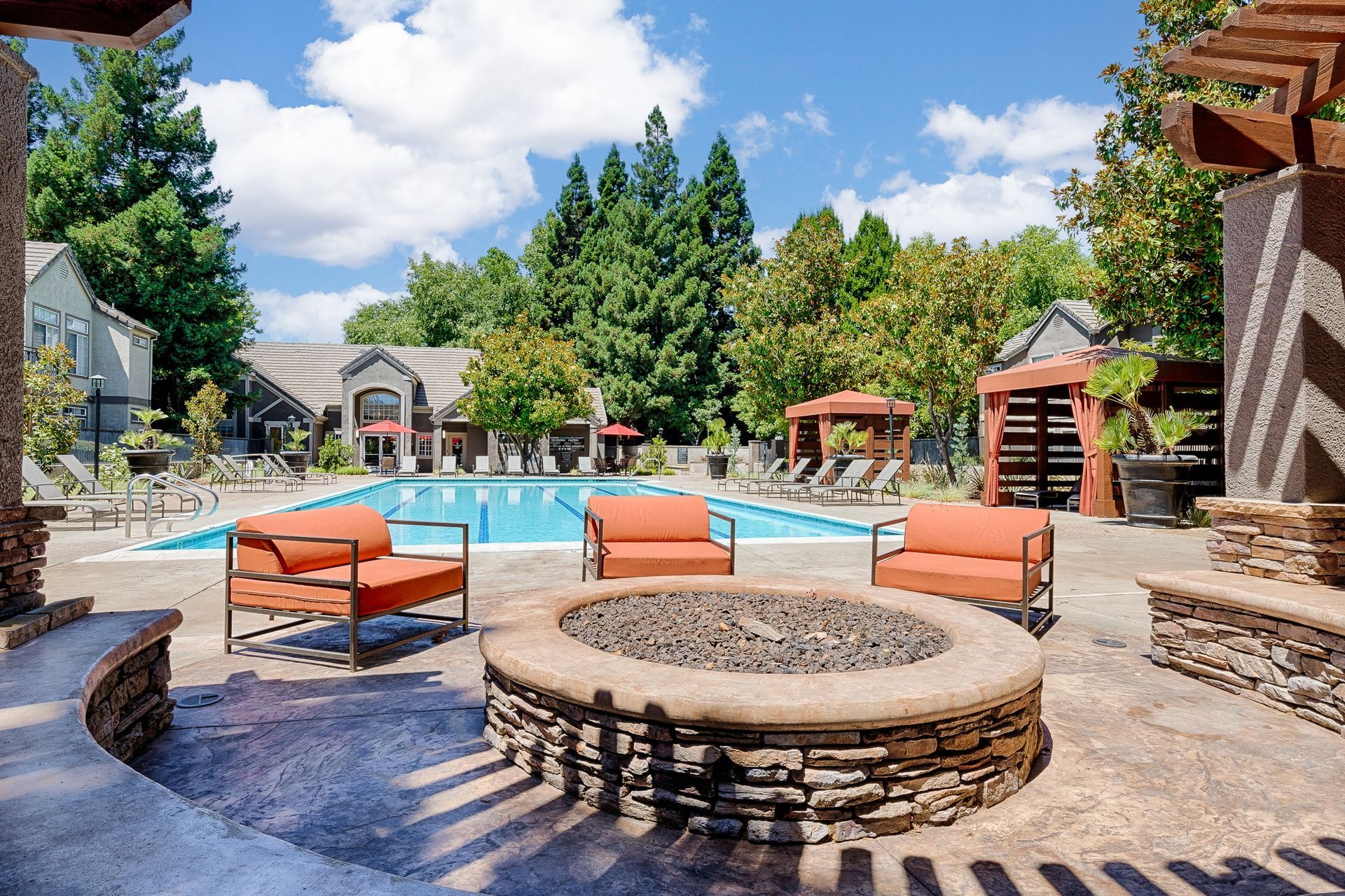 Outdoor pool area with lounge chairs, cabanas, and a circular fire pit in a landscaped courtyard.