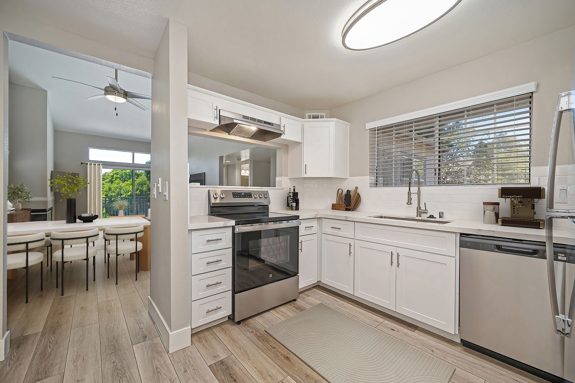 Bright white kitchen with black appliances, wooden floors, and a dining area visible through the doorway