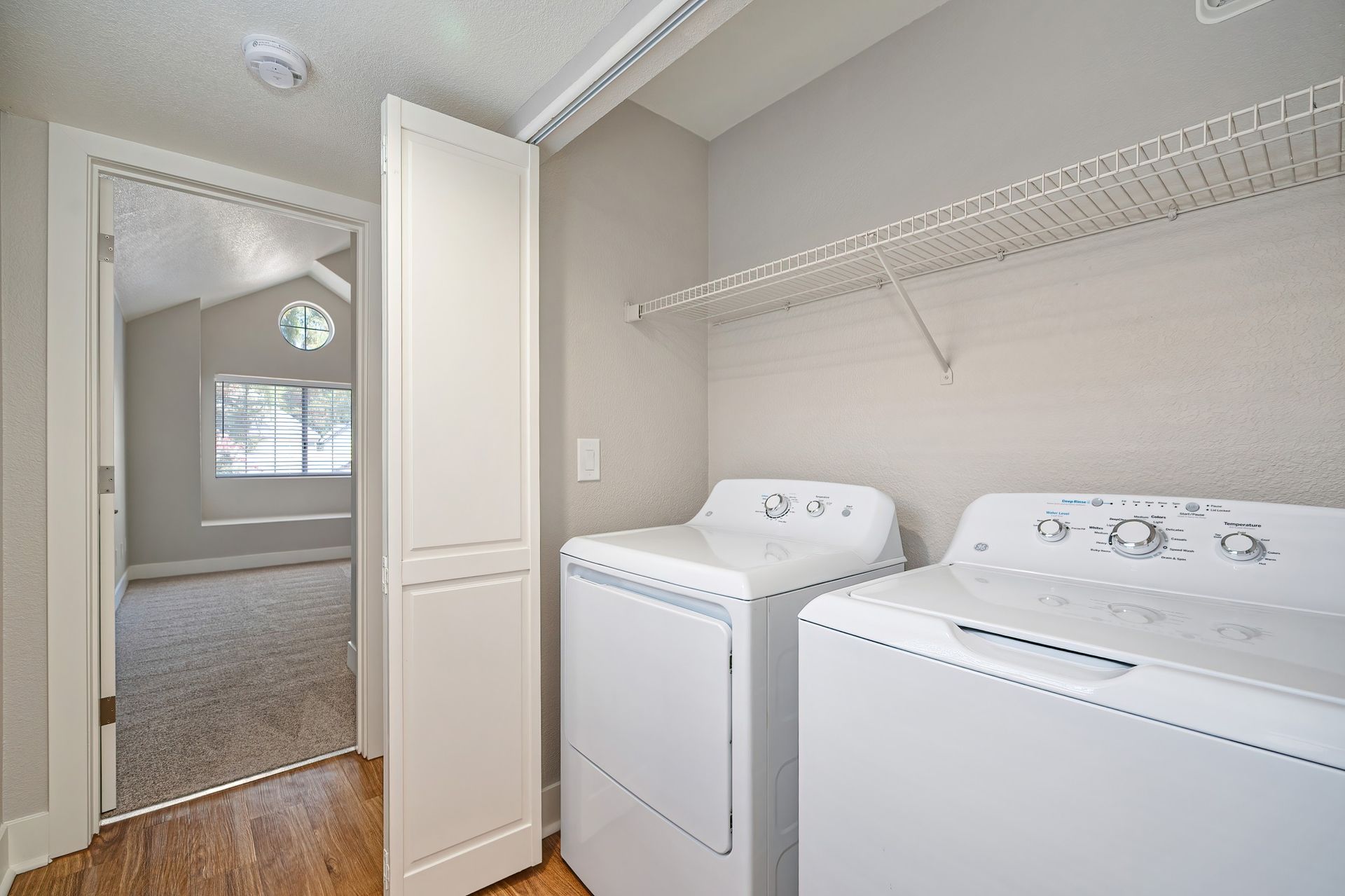 Laundry room with white washer and dryer, open doorway, and wire shelf on the wall.