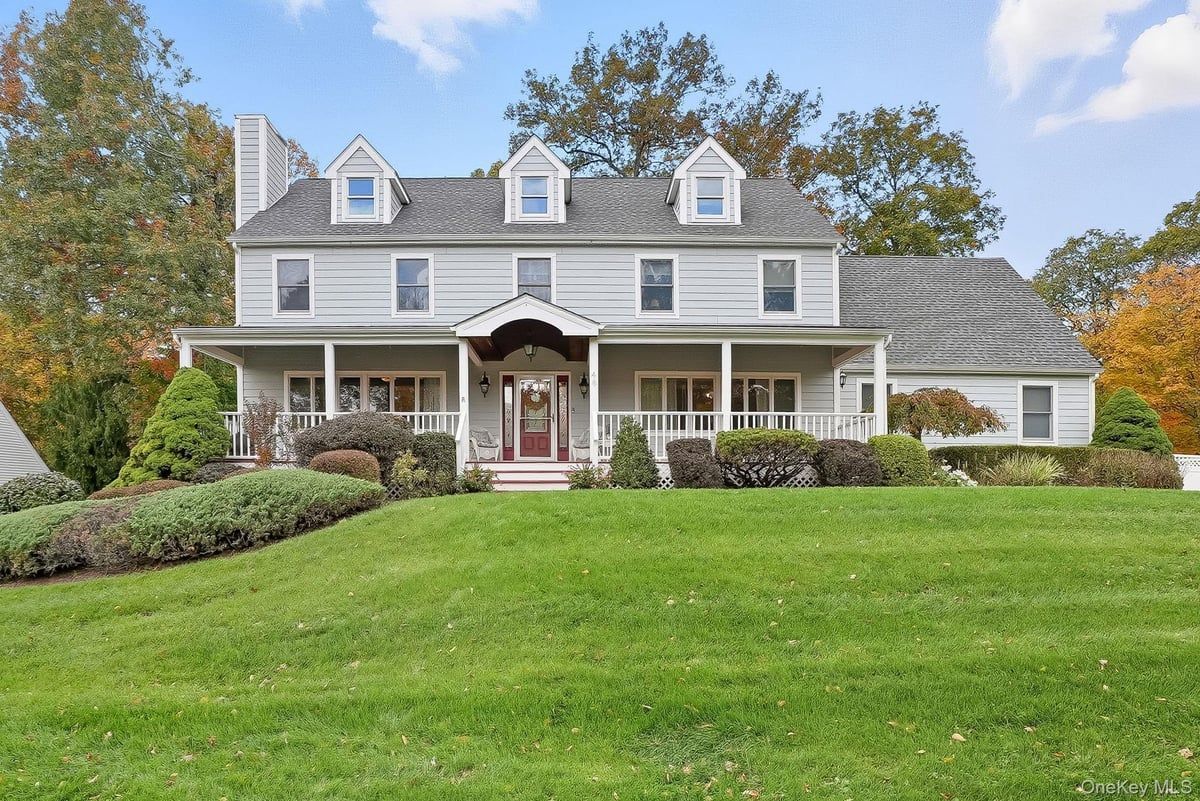 Large gray suburban house with dormer windows, a front porch, and a wide green lawn under a blue sky