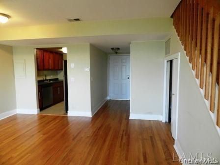 Empty living room with hardwood floors, white walls, a kitchen pass-through, and stairs on the right.