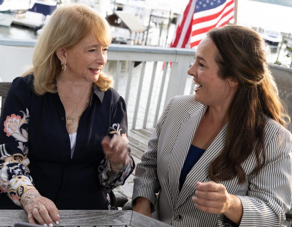 Two women smiling and talking at a table outdoors, with an American flag in the background