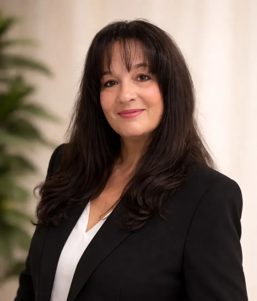 Woman in a black blazer and white blouse, smiling against a soft beige background