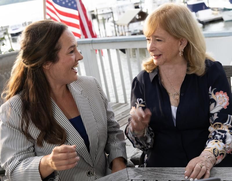Two women smiling and talking at a table, with an American flag in the background.