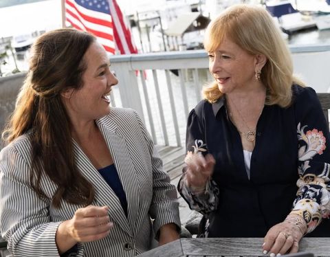 Two women smiling and talking at a table, with an American flag in the background