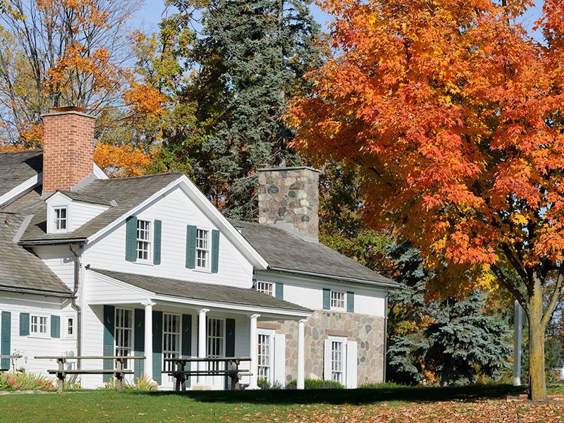 White house with porch and stone chimney amid autumn trees with orange and yellow leaves