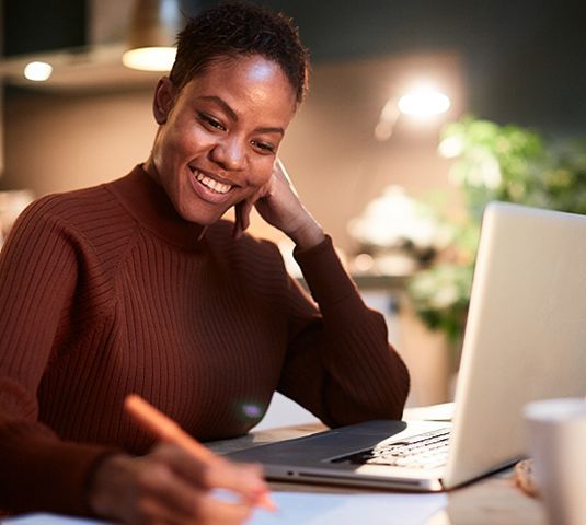 Smiling person working on a laptop at a desk, writing in a notebook in a warm indoor setting