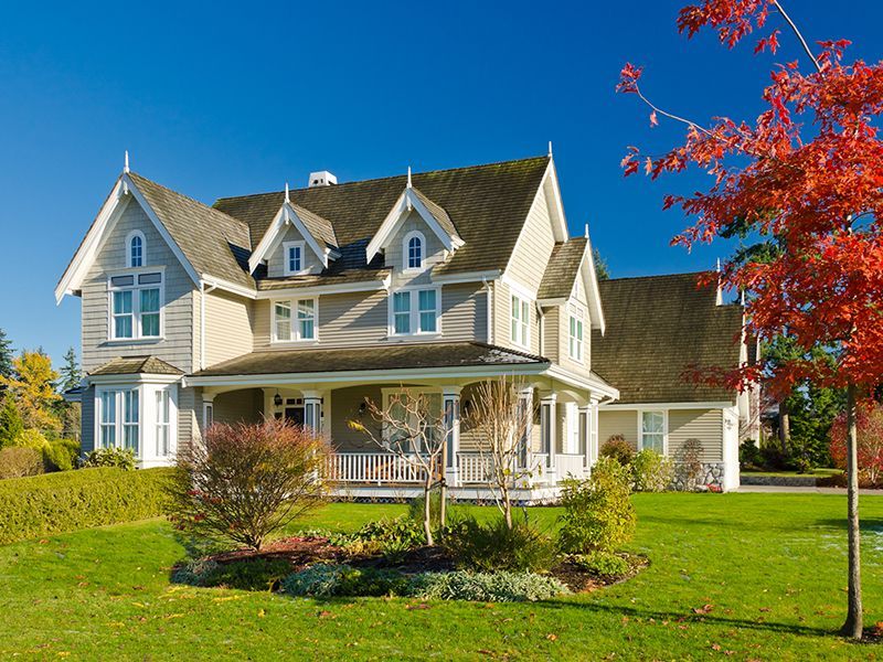 Large light-gray house with white trim, wraparound porch, and colorful autumn trees under a clear blue sky
