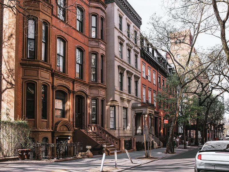 Tree-lined city street with historic brownstone buildings and parked cars on an overcast day