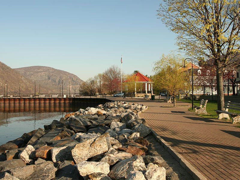 Riverside walkway with rocks, benches, trees, and a red-roof gazebo on a sunny day