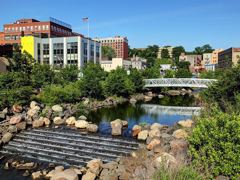 River with small waterfall, rocks, trees, and downtown buildings under a clear sky
