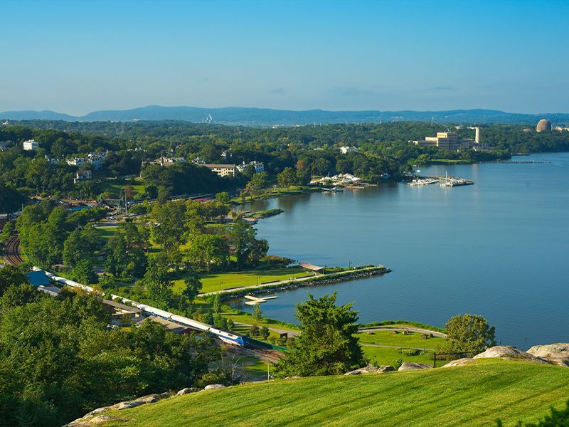 Green hillside overlooking a blue bay, with trees, shoreline, and distant buildings under a clear sky