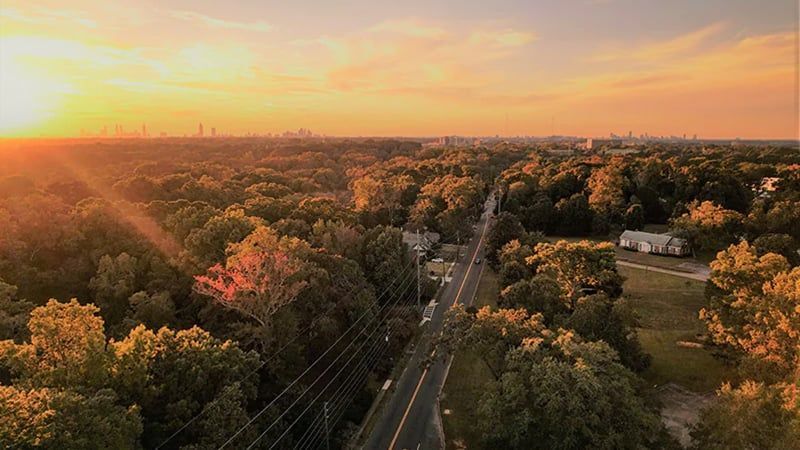 Sunset over a tree-lined suburban road, with warm orange sky and distant city skyline.
