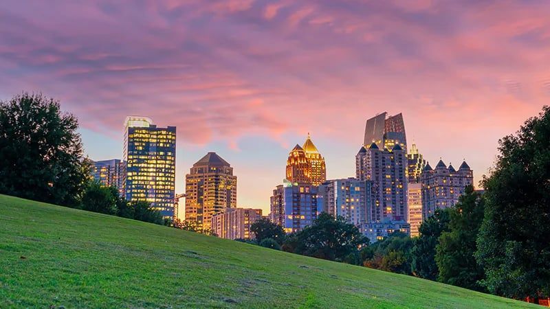 City skyline at sunset above a green grassy hill under a pink-orange sky