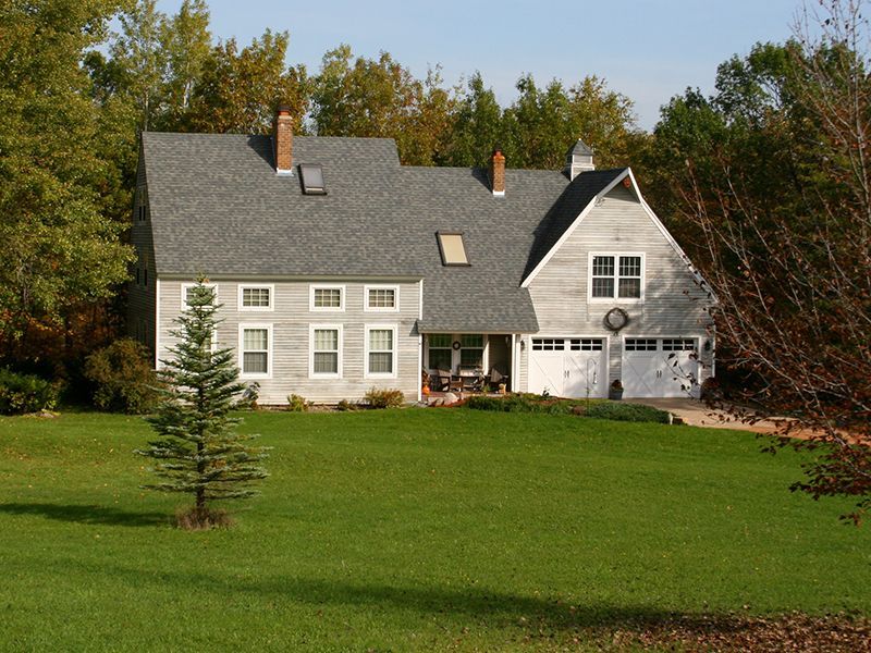 White house with gray roof and garage on a green lawn, surrounded by trees under clear sky