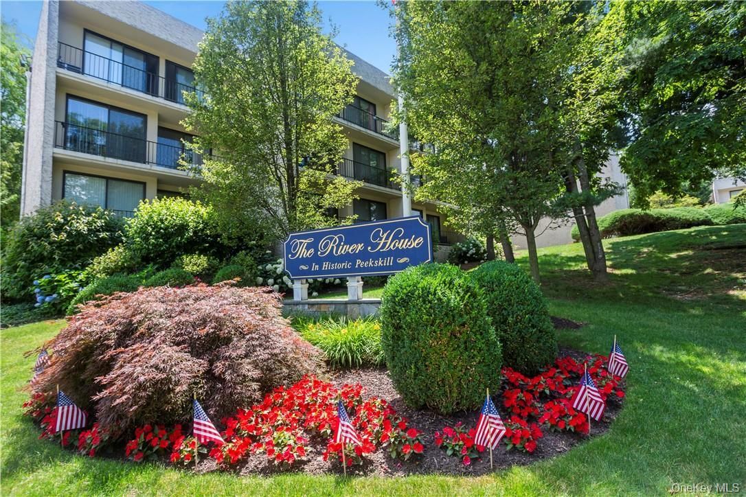 Apartment building entrance with manicured shrubs, red flowers, and a blue sign in a sunny garden setting