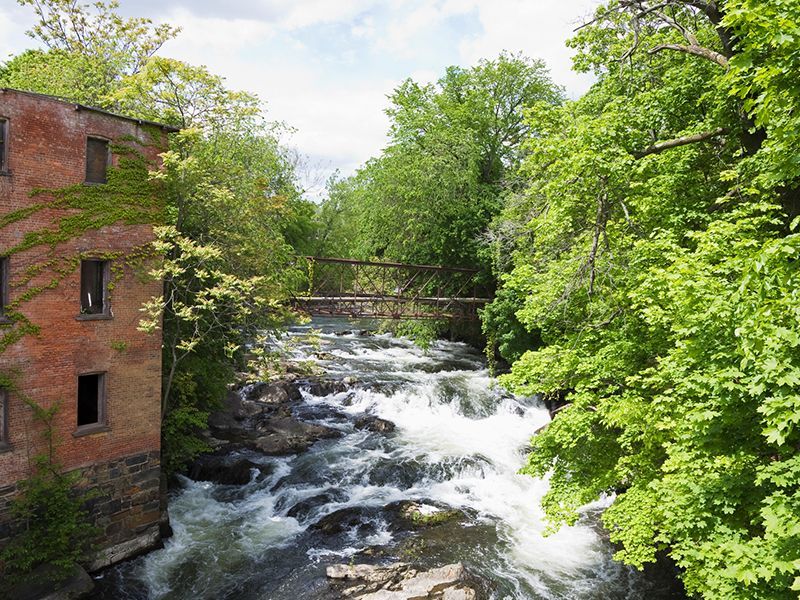 Rushing river beside a red brick building, with a small bridge and green trees along the bank