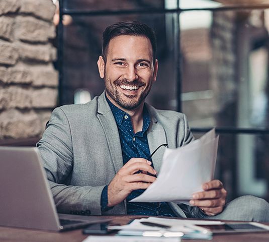 Smiling man in a gray blazer reviewing papers at a laptop in a cozy cafe