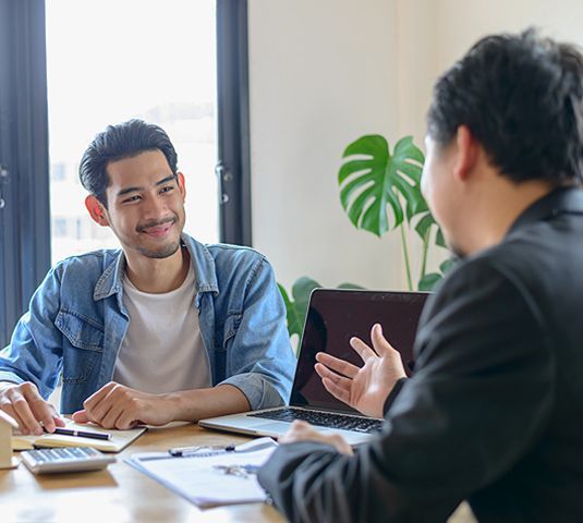 Two people talking at a desk in a bright office, with papers, a laptop, and a potted plant.