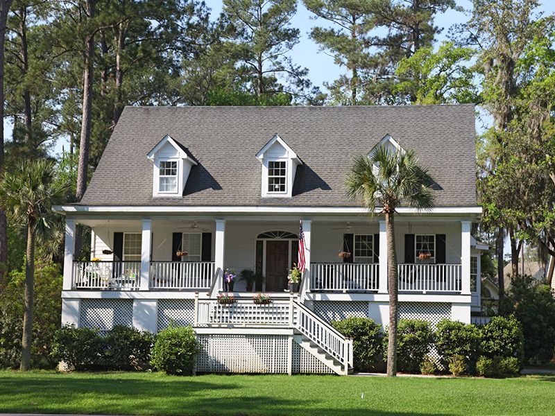 White two-story house with wraparound porch, dark roof, dormer windows, and front steps amid trees and lawn