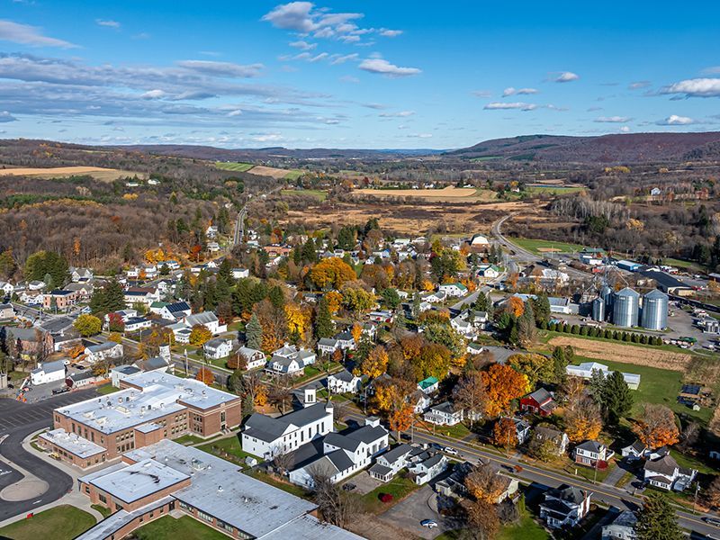 Aerial view of a small autumn town with houses, trees, and rolling hills under a blue sky