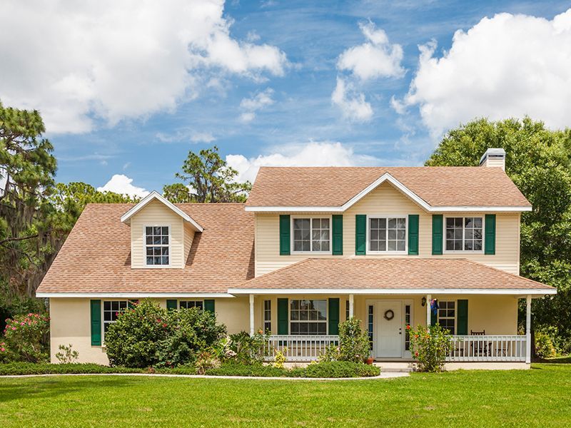 Two-story beige house with green shutters, brown roof, and white porch under a blue sky