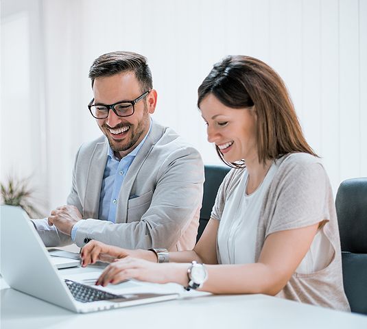 Two coworkers smiling and typing on laptops at a bright office desk