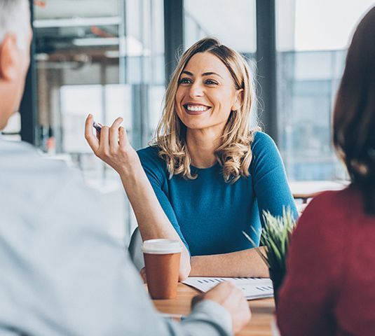 Smiling woman in a blue top speaking at a meeting table with coffee cups and colleagues nearby