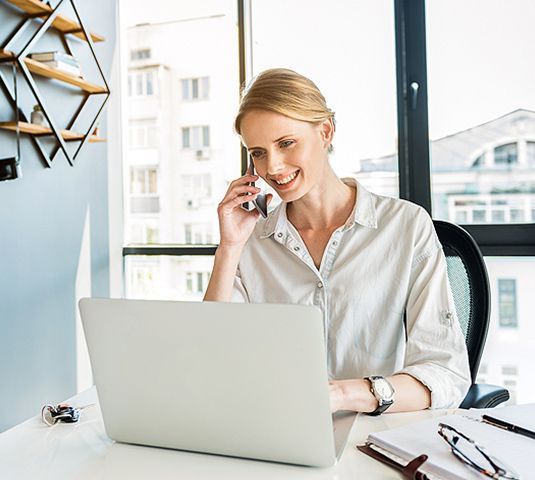 Person smiling and talking on the phone at a laptop in a bright office