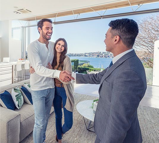 Two men shaking hands in a bright modern living room, with a woman smiling nearby and water views outside.