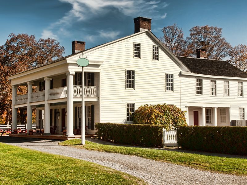 White colonial-style house with porch, hedge, and gravel driveway under autumn trees